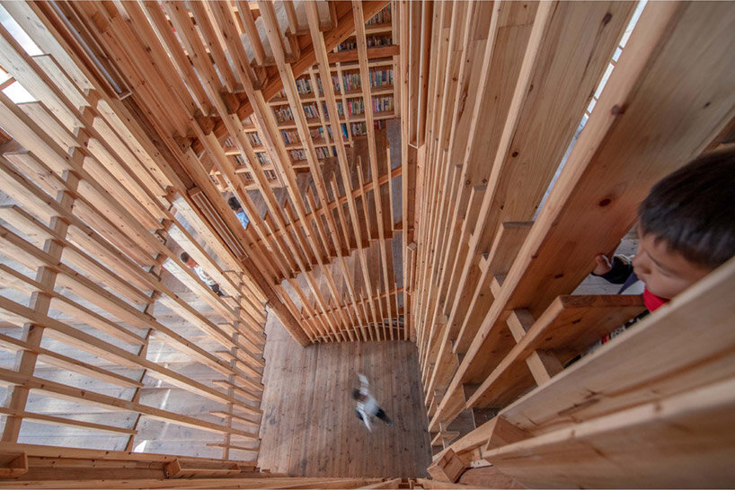 matriz-estantes-compoe-fachada-luminosa-china-sai-zhao-biblioteca-escada-construcao-madeira vista do 'miolo' da escada em biblioteca comunitária de madeira, com duas crianças circulando em diferentes pavimentos.