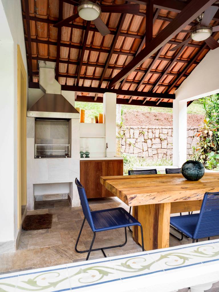 15-retrofit-em-duas-casas-de-familia-recupera-estilo-colonial-das-construcoes Varanda com mesa de madeira, churrasqueira branca e cadeiras azuis