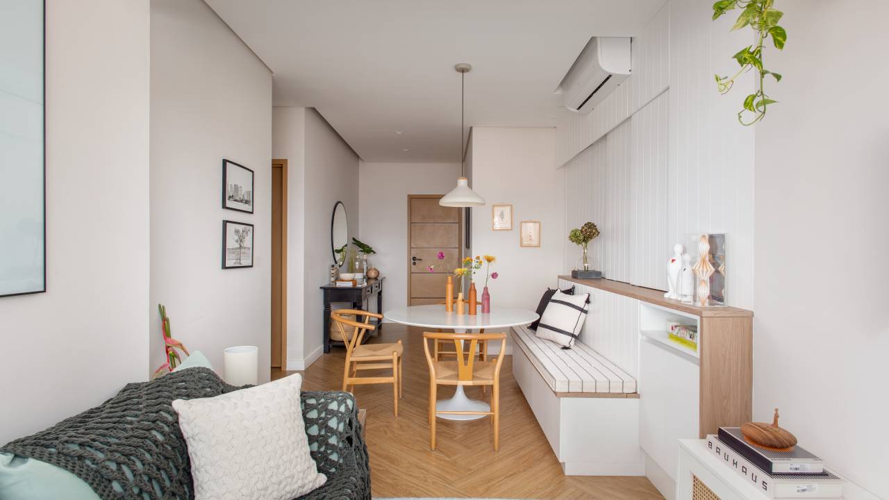 Delicadeza: apê de 66 m² tem cozinha aberta e cabeceira com bordados. Projeto de Memoá Arquitetos. Na foto, sala de estar pequena, banco fixo, ar condicionado.