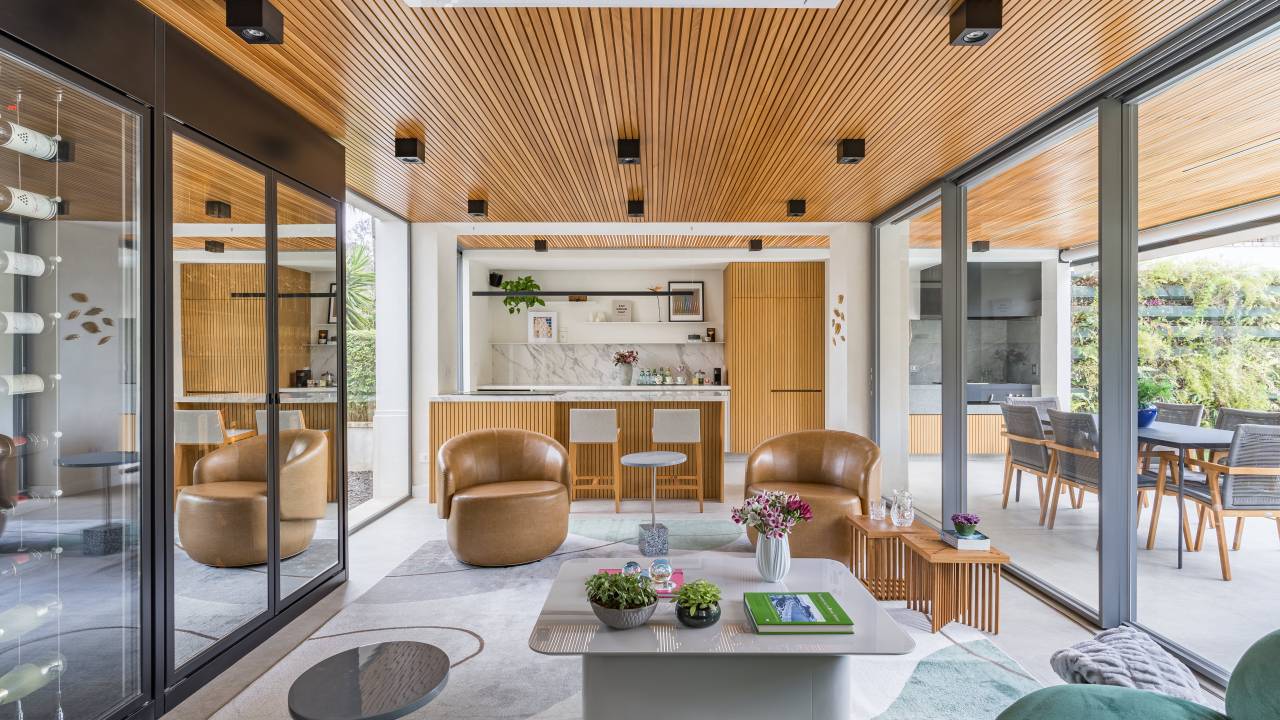 Adega-cristaleira funciona como divisória em área social de casa. Projeto de Spaço Interior. Na foto, sala de estar, teto ripado, poltrona de couro caramelo.
